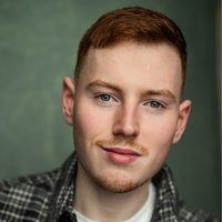 Actor Nicholas Marshall, smiles at the camera. He is in front of a green background - making his red hair shine.