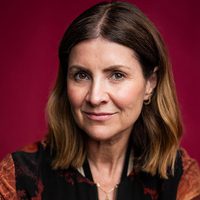 Actor Jayne McKenna smiles at the camera she sits in front of a red background and wears an elegantly printed top to match.