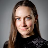 Naomi Stirrat smiles at the camera, she has long hair, wears a black high-necked top and has piercing grey eyes.