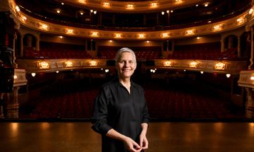 Jane Speirs stands centre stage with a theatre auditorium in the background.