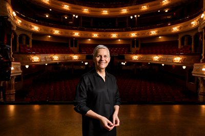 Jane Speirs stands centre stage with a theatre auditorium in the background.