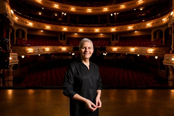 Jane Speirs stands centre stage with a theatre auditorium in the background.