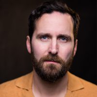 Robert Jack sports a serious expression yet he remains warm. He wears an orange shirt.