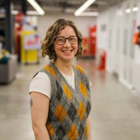 Becca stands smiling in a hallway. She wears an argyle vest and glasses.
