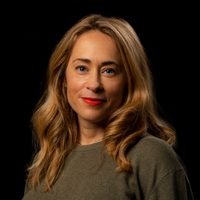 Catherine smiles at the camera, she wears bright red lipstick and a dark grey jumper.