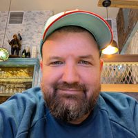 Danny smiles at the camera. He wears a baseball cap and blue top. He has a brown beard.