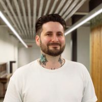 Derek smiles at the camera, he wears a white t-shirt and has lots of tattoos.