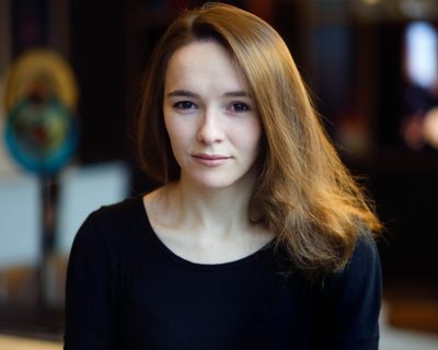 Emily looks intently at the camera . She wears a black top and has honey brown hair
