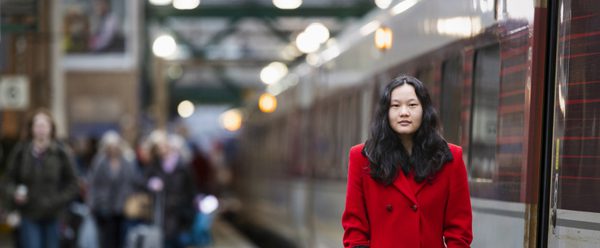 Gemma stands on a platform at a railway station. She wears a striking red coat, a train stands beside her.