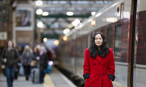 Gemma stands on a platform at a railway station. She wears a striking red coat, a train stands beside her.