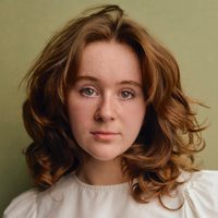 Holly is a young woman in her early twenties. She wears a white top, has hazel-green eyes, and light brown hair. She stands in front of a pistachio coloured background.