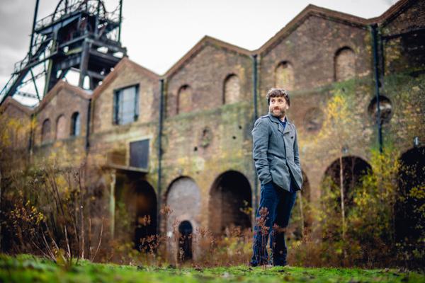 A man stands outside the National Mining Museum, a red brick building with mining machinery attached. He is looking to his right.