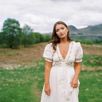 Mairi wears a white dress and stand in a meadow.