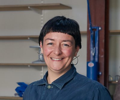 Headshot of Nic Green, a photo of a woman with cropped black hair, silver hoop earrings and a blue shirt.