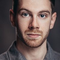 Robin looks at the camera, he wears a grey shirt and the background is also grey