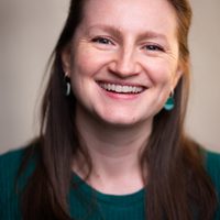 Shonagh smiles brightly at the camera, she wears a dark green jumper with matching earrings. She has brown hair.