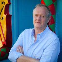 Tony has his arms folded and leans against the wall with a yellow cello in the background.