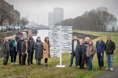 A group of people stand on the banks of a ca nal next to a sign post with various signs pointing towards locations across Scotland.