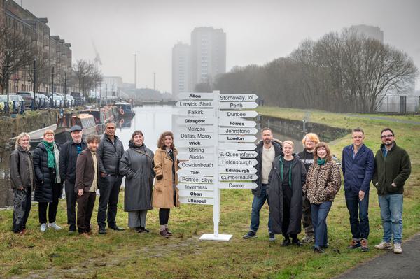 A group of people stand on the banks of a ca nal next to a sign post with various signs pointing towards locations across Scotland.