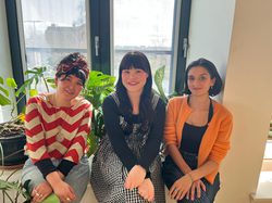 The three fellows, Andrea Ling, Hannah Low, and Leah Byrne, sit in front of a window and smile happily at the camera