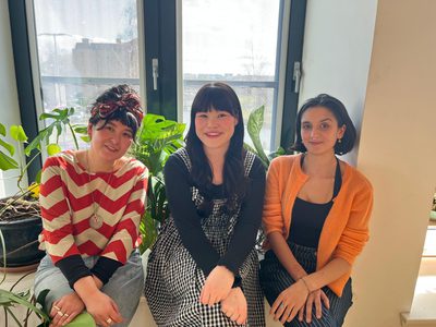 The three fellows, Andrea Ling, Hannah Low, and Leah Byrne, sit in front of a window and smile happily at the camera