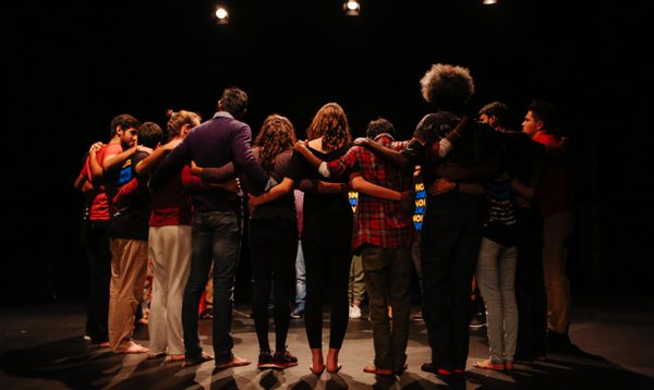 Actors stand on a stage with their arms around each others shoulders