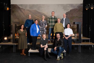 A group of people stand on platforms, in front of a painted backdrop of the Scottish Highlands, with large black brackets on either side and stage lights.