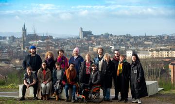A group of artists stood at the viewing point at the Glasgow Claypits, with a blue sky and the Glasgow skyline behind them.
