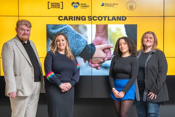 Alt text: Four people stand in front of an orange screen displaying the Caring Scotland title and a photo of a child holding an adult's hand. They are all looking at the camera.