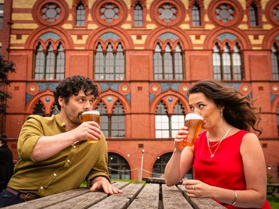 A man and woman sit at a picnic bench, they make eye contact as they sip pints