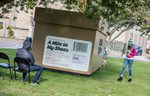 Large cube-shaped installation labelled “A Mile in My Shoes” sits on a grassy area under trees. Two people engage with the installation -- one seated on a chair facing the structure and another standing beside it.
