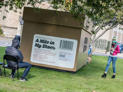 Large cube-shaped installation labelled “A Mile in My Shoes” sits on a grassy area under trees. Two people engage with the installation -- one seated on a chair facing the structure and another standing beside it.