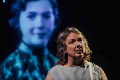 A woman with shoulder length grey hair in a grey dress is facing forward. Behind her, the face of a younger person is projected behind her.
