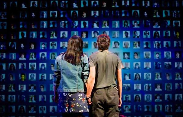 Adam holds his wife's hand and looks at a wall of faces on a big screen.