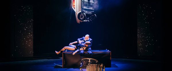 A man in a suit sits centre stage holding a crash test dummy. A car is suspended over his head.