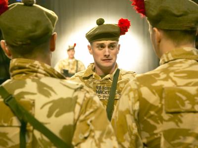 An actor in military attire stands pulling a puzzled face at two other men