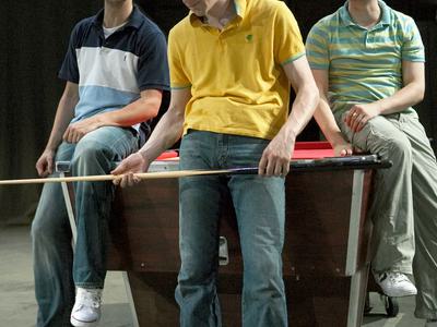 Three men in casual clothing pose in front of a pool table. The centre man holds a pool cue