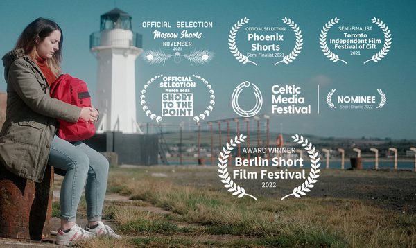 a woman sits holding a red backpack. in the distance behind her is a white lighthouse. The film's various awards have been overlaid on the right side of the image.
