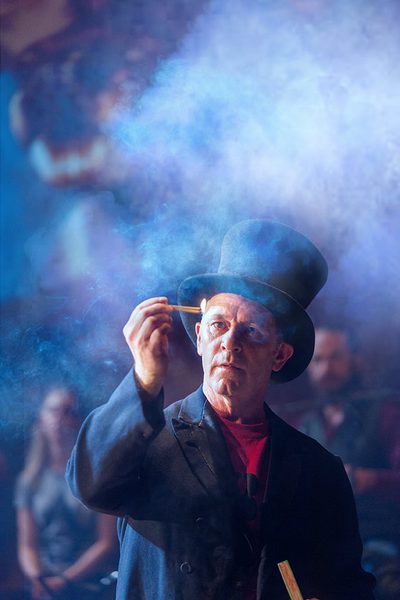 An older man wearing a top hat stares at a lit match he holds in his hand, a look of determination on his face. He is surrounded by smoke and we can see a crowd gathered in the background.