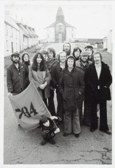 A black and white photo of a group of actors stand in the middle of the street in front of the Round Church in Bowmore. At the front of the group, a child is holding a flag that reads ‘7:84’