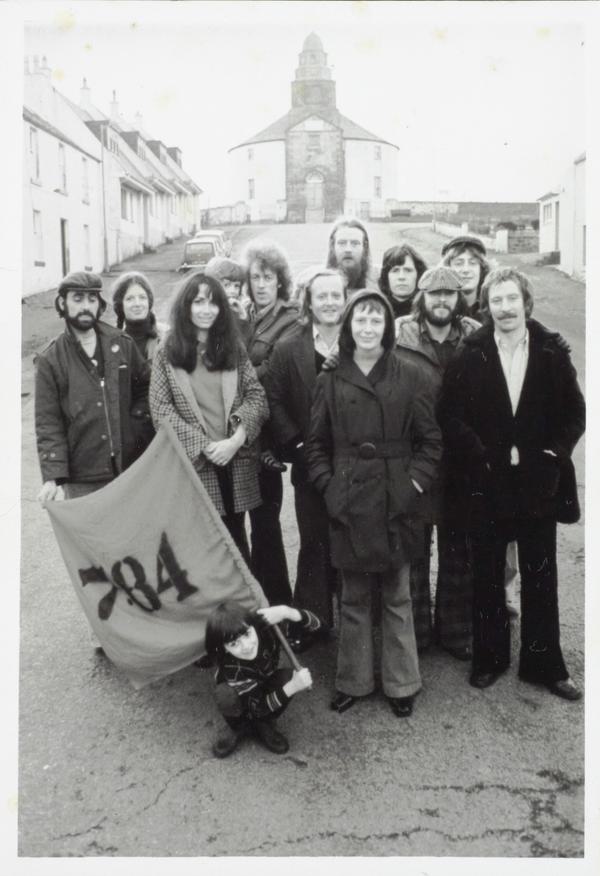 A black and white photo of a group of actors stand in the middle of the street in front of the Round Church in Bowmore. At the front of the group, a child is holding a flag that reads ‘7:84’