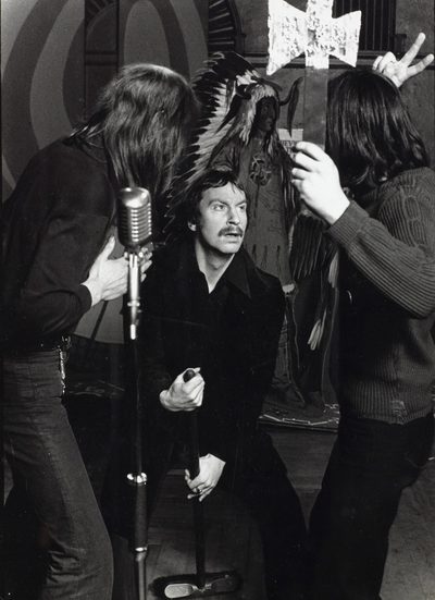 A black and white photo of a man holding a broom. There are two people on either side of him, one is holding a pretend axe. The man in the middle is looking up at them.