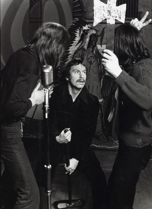 A black and white photo of a man holding a broom. There are two people on either side of him, one is holding a pretend axe. The man in the middle is looking up at them.
