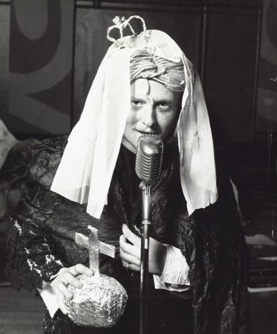 A black and white photo of an actor dressed as Queen Victoria, with a string wig, a small paper crown and a tinfoil globus cruciger.