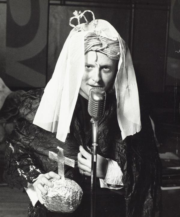 A black and white photo of an actor dressed as Queen Victoria, with a string wig, a small paper crown and a tinfoil globus cruciger.