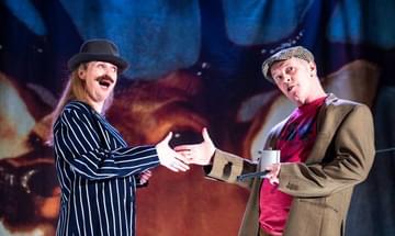 Two performers are about to shake hands. Jo Freer, to the left, wears a fake moustache, a stripy blue blazer, and a small bowler hat. Billy Mack, to the right, wears a green woollen hat, a green woollen blazer and a red tshirt. The Monarch Of The Glen painting, featuring a stag, is visible as a backdrop.