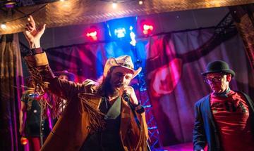 A performer wearing a fringed jacket and a hat is singing into a microphone