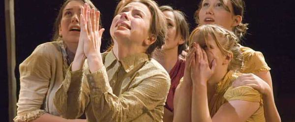 Five young girls look up, scared. Some clasp their hands over their mouths, others hold on to each other. In the foreground on had her palms pressed together in a praying gesture.