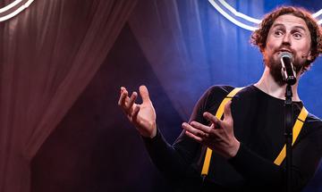 A man stands at a microphone, he wears a black top with yellow suspenders. Behind him are curtains and a neon light.