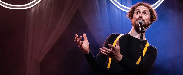 A man stands at a microphone, he wears a black top with yellow suspenders. Behind him are curtains and a neon light.